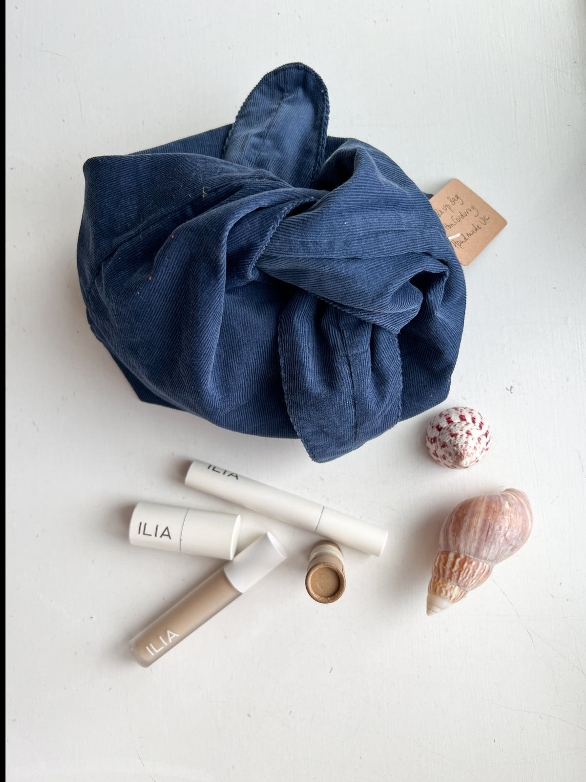 A dark indigo blue fabric pouch, tied in a knot, displayed on a white surface alongside cosmetic products by ILIA and decorative seashells.